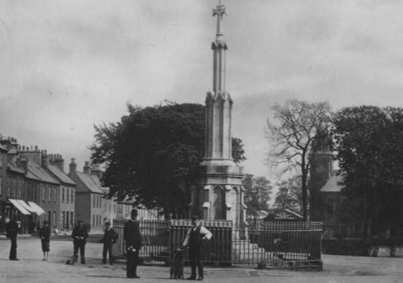 Old photograph, dated around 1900, showing the Mercat Cross in Wigtown. Some figures are standing around the monument, dressed in period clothing.